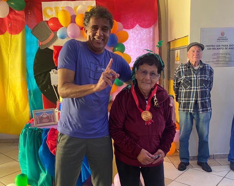 Na imagem, duas pessoas em foco. À esquerda, um homem de camiseta azul e calças pretas, sorrindo, e fazendo o símbolo do número um com a mão. Ao seu lado, uma senhora que veste blusa vinho e calças pretas, com uma medalha em seu pescoço e segurando notas de 